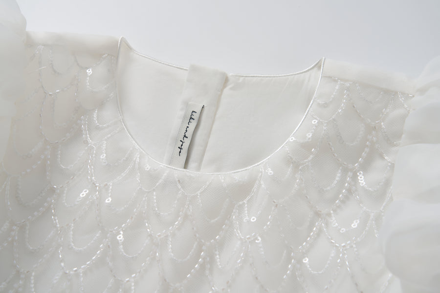 Close-up of the Ballerina Whisper Dress by KIKI and JOJO, showcasing sequin scallop patterns and beaded embroidery, with a back button closure and an inner collar fabric label.