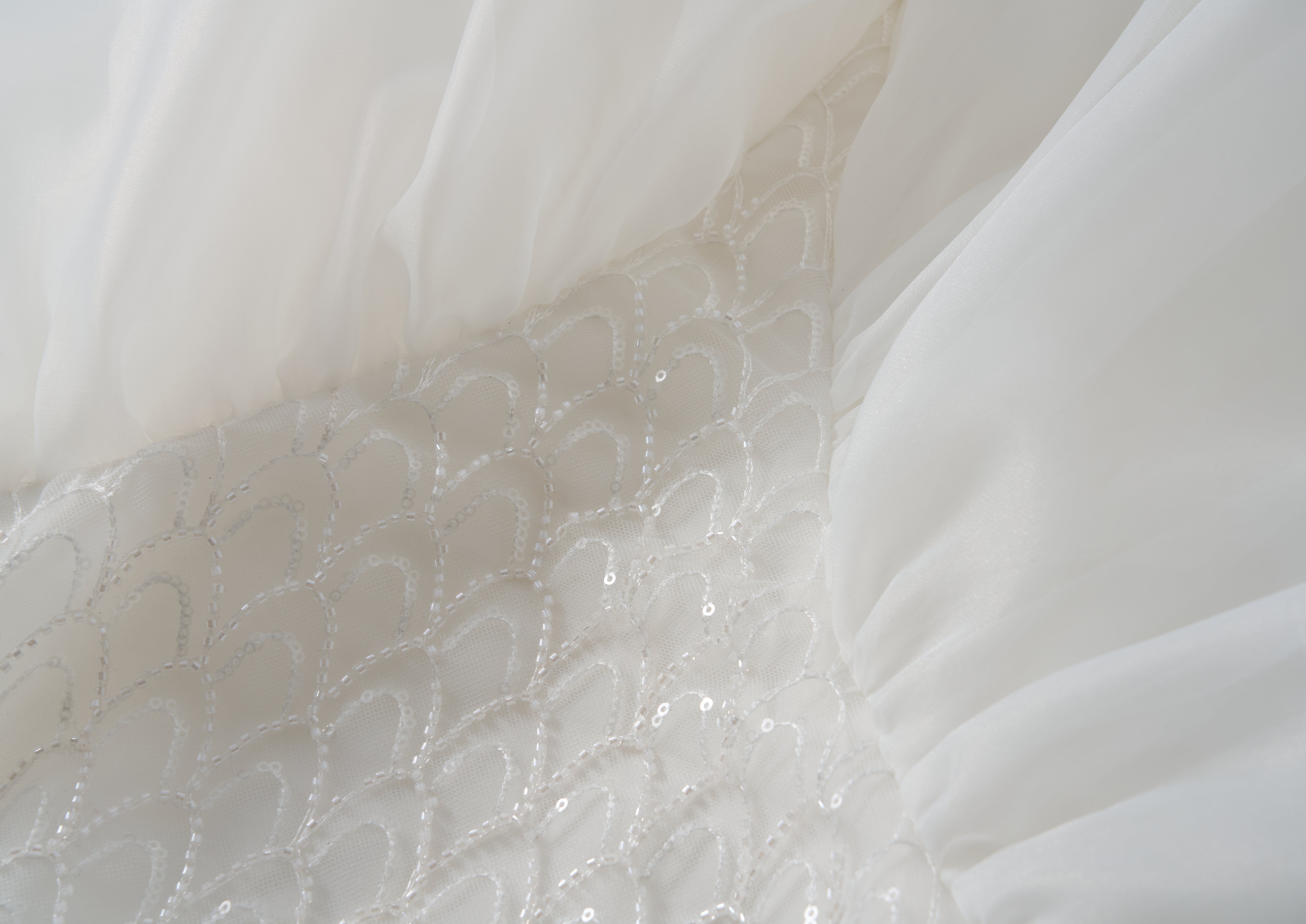 Close-up of the Ballerina Whisper Dress by KIKI and JOJO, featuring white fabric with sheer gathered tulle sides and a central panel adorned with a scalloped sequin pattern, evoking an elegant ballet-inspired design.