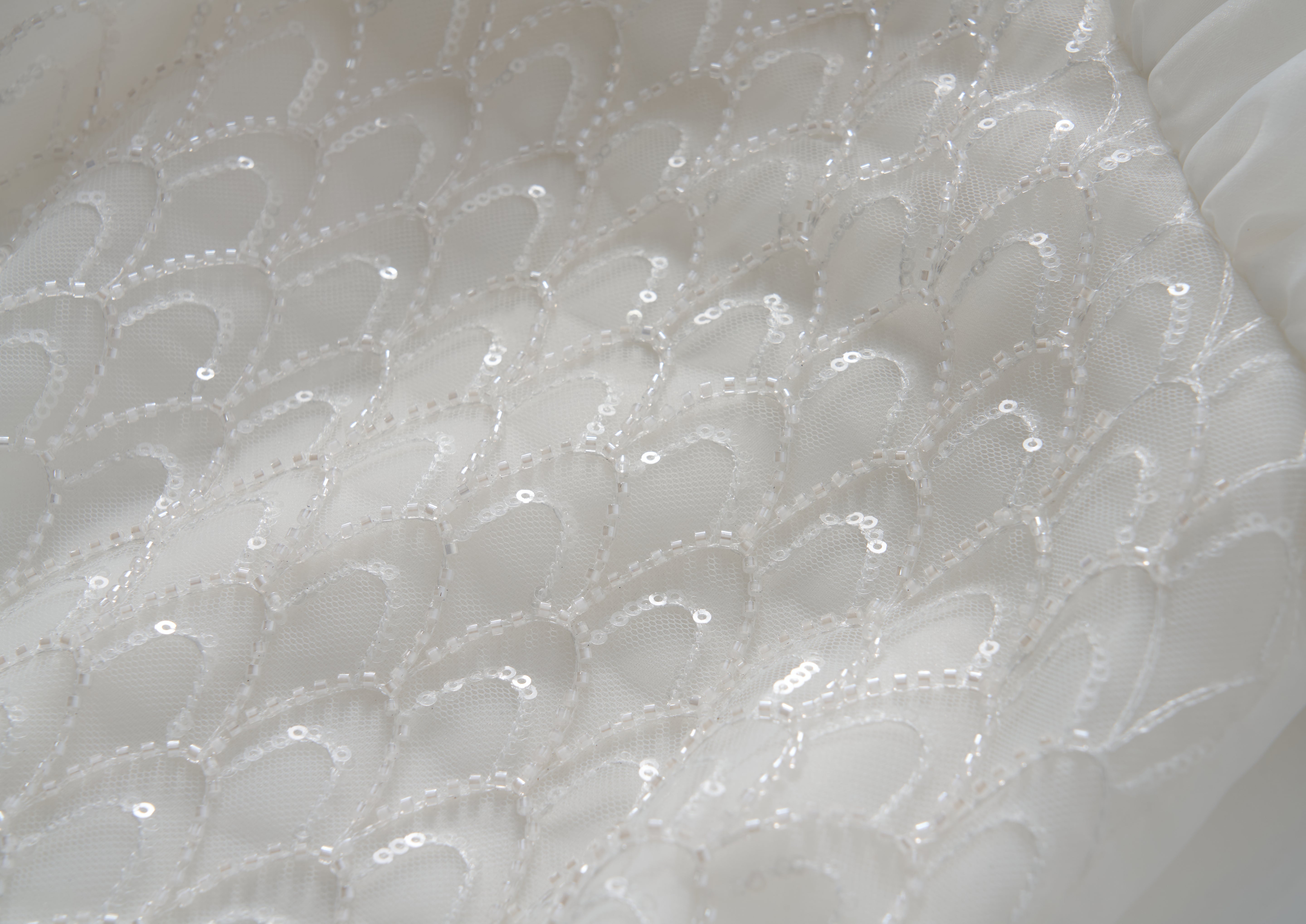 Close-up of white tulle fabric from the Ballerina Whisper Dress by KIKI and JOJO, featuring a repeating scalloped pattern with sequins and beaded embroidery for a textured, shimmering effect.