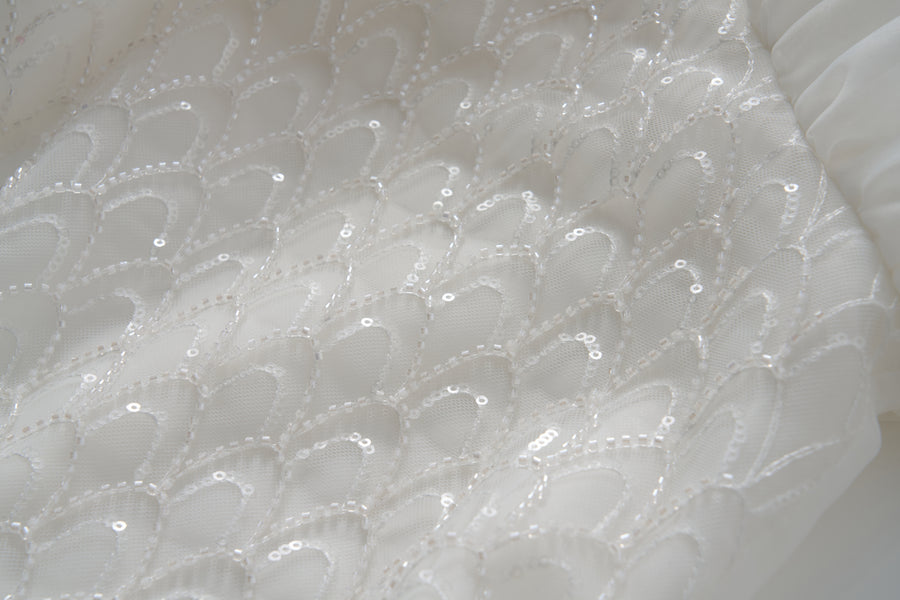 Close-up of white tulle fabric from the Ballerina Whisper Dress by KIKI and JOJO, featuring a repeating scalloped pattern with sequins and beaded embroidery for a textured, shimmering effect.
