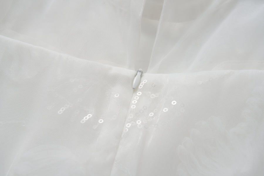 Close-up of the Whisper Bloom Dress by KIKI and JOJO—a white mini dress with floral embroidery, zipper closure, sheer fabric adorned with subtle sequins, and delicate bead accents.