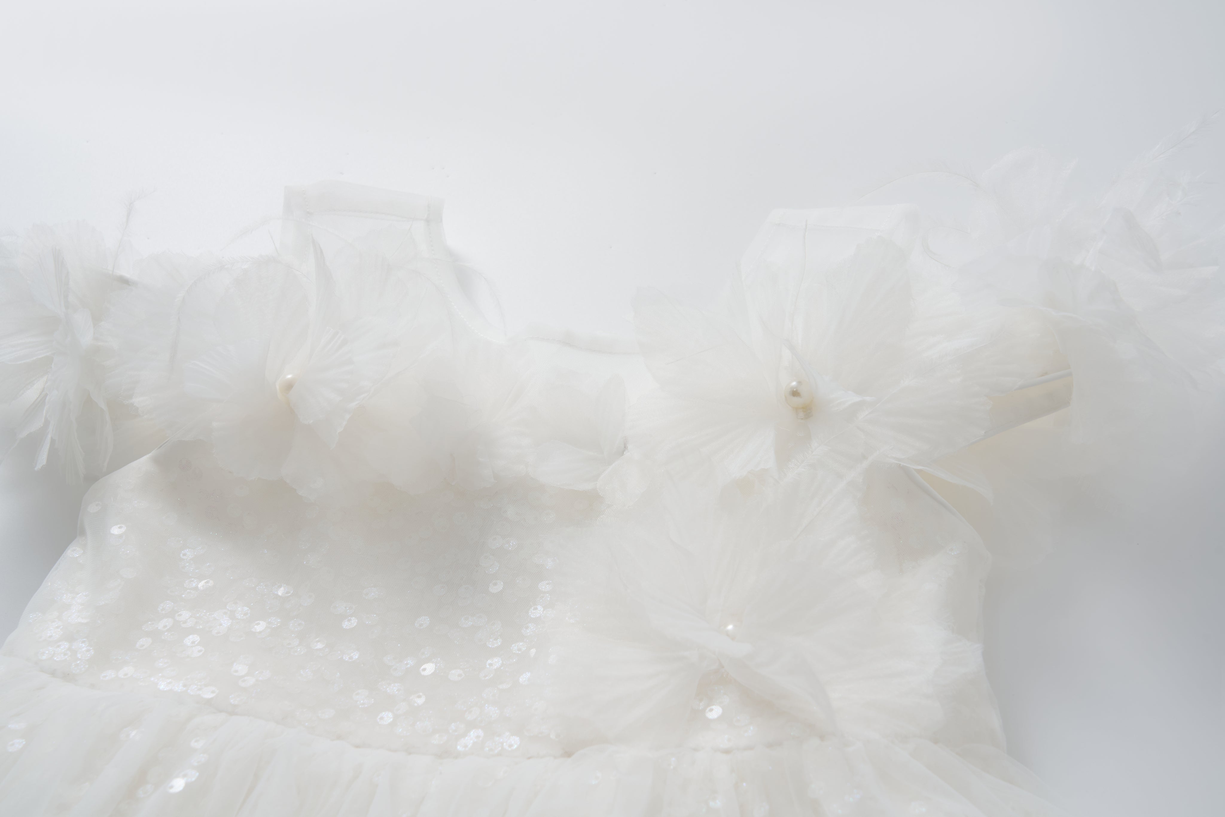 The Feathered Whisper Dress by KIKI and JOJO features white satin with sequin details and large flower appliques on the shoulders, styled in an elegant off-shoulder A-line silhouette and photographed against a plain white background.