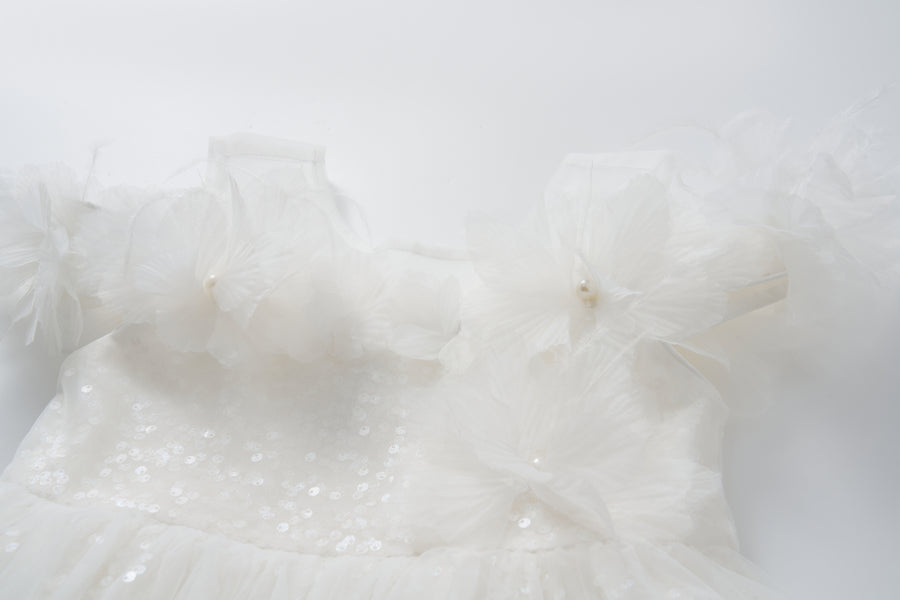 The Feathered Whisper Dress by KIKI and JOJO features white satin with sequin details and large flower appliques on the shoulders, styled in an elegant off-shoulder A-line silhouette and photographed against a plain white background.