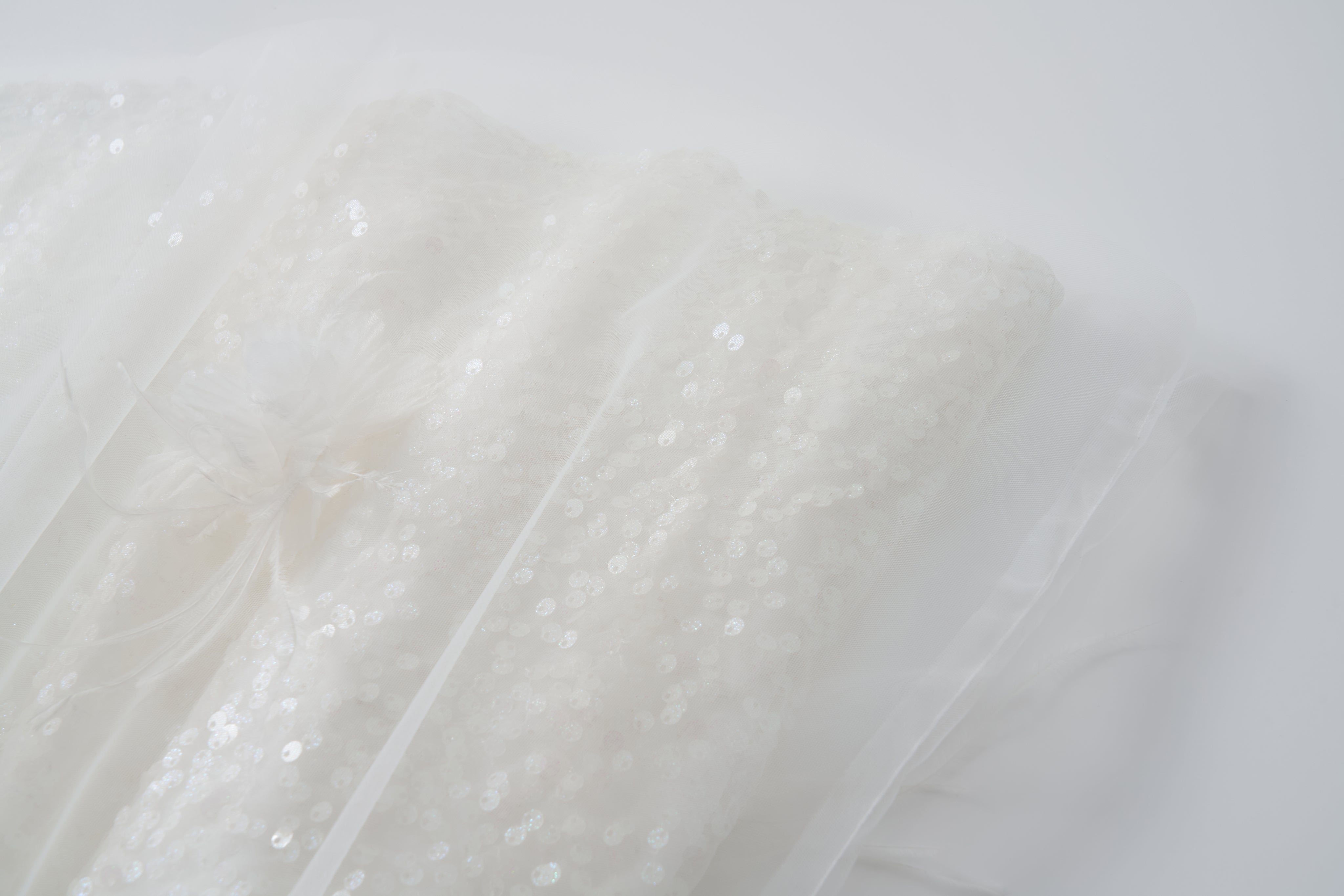 Close-up of the Feathered Whisper Dress by KIKI and JOJO—white sequin fabric with a sheer overlay, off-shoulder A-line design, and a delicate decorative bow on a softly lit neutral background.