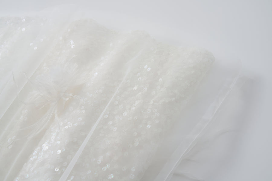 Close-up of the Feathered Whisper Dress by KIKI and JOJO—white sequin fabric with a sheer overlay, off-shoulder A-line design, and a delicate decorative bow on a softly lit neutral background.