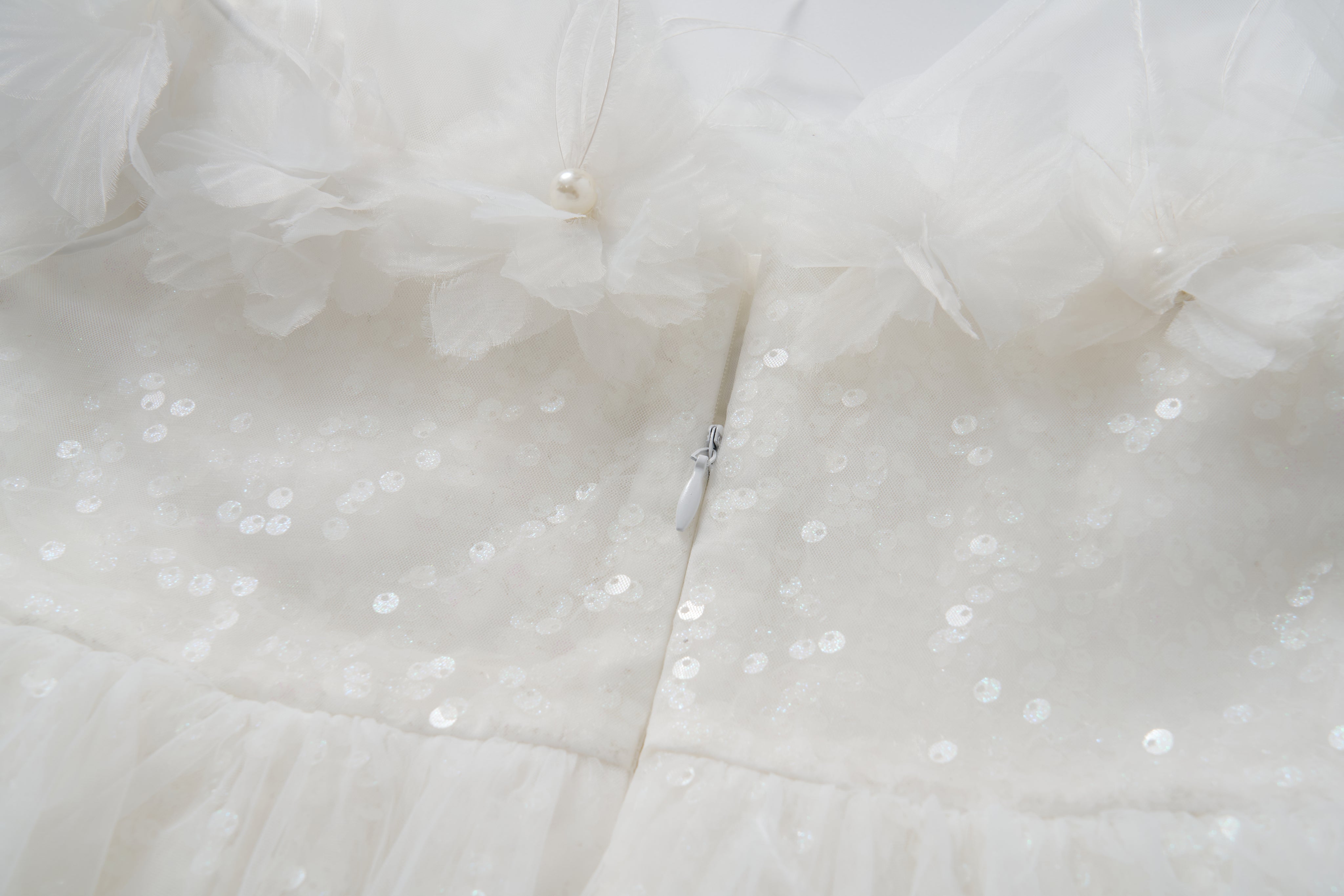 Close-up of the Feathered Whisper Dress by KIKI and JOJO—a white sequined fabric with floral tulle, pearl accents, and a hidden center zipper—ideal for an off-shoulder A-line silhouette.