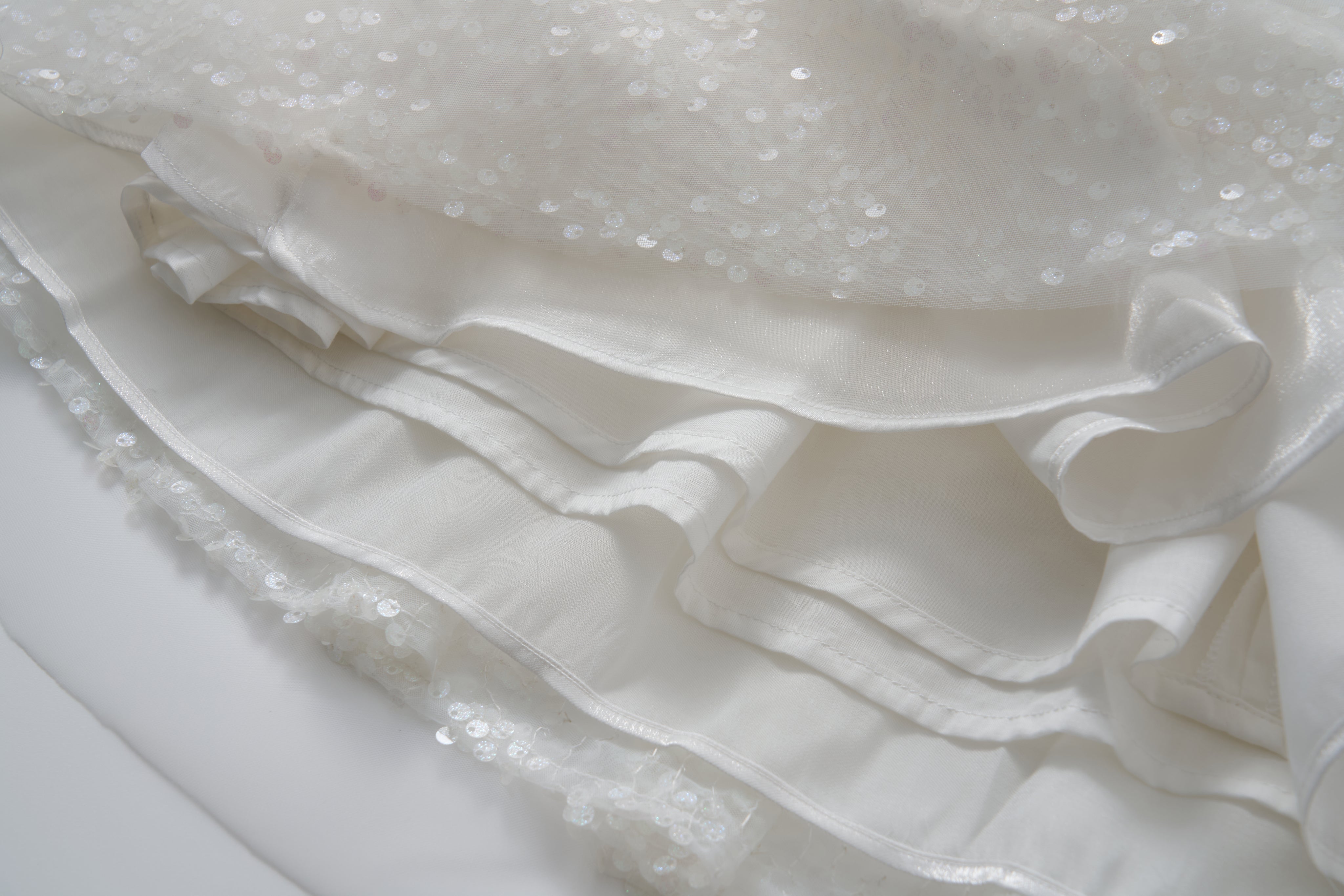 Close-up of the Feathered Whisper Dress by KIKI and JOJO, featuring layered white fabric, sequins, ruffles, and delicate floral feather embellishments that highlight its exquisite texture and decorative details.