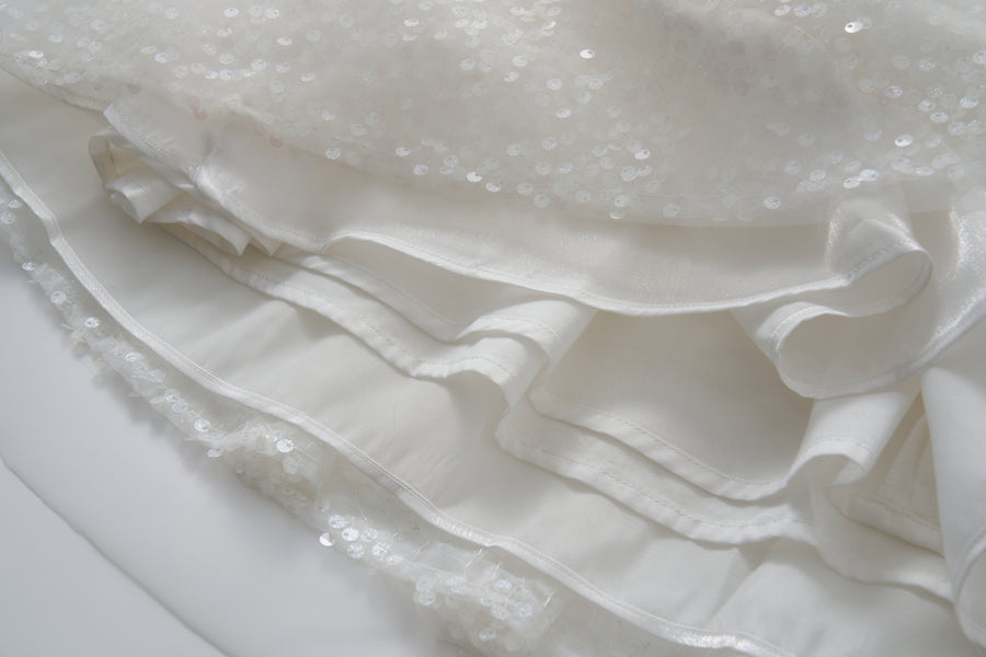 Close-up of the Feathered Whisper Dress by KIKI and JOJO, featuring layered white fabric, sequins, ruffles, and delicate floral feather embellishments that highlight its exquisite texture and decorative details.
