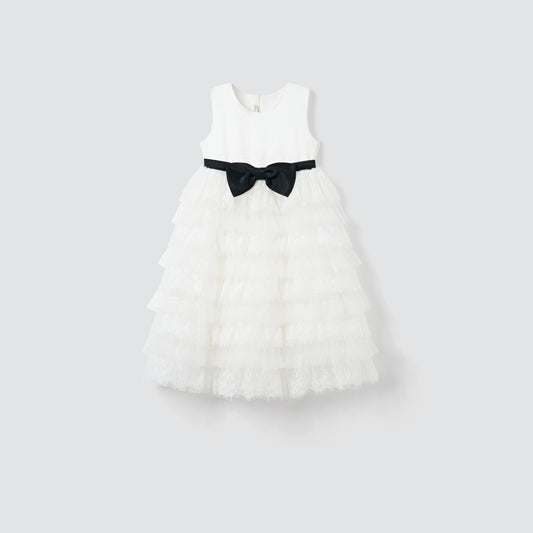 The Monochrome Whisper Dress by KIKI and JOJO is a sleeveless white girl's dress featuring lace ruffles, a black ribbon waist bow, and a tiered tulle cake skirt, shown against a plain light background.