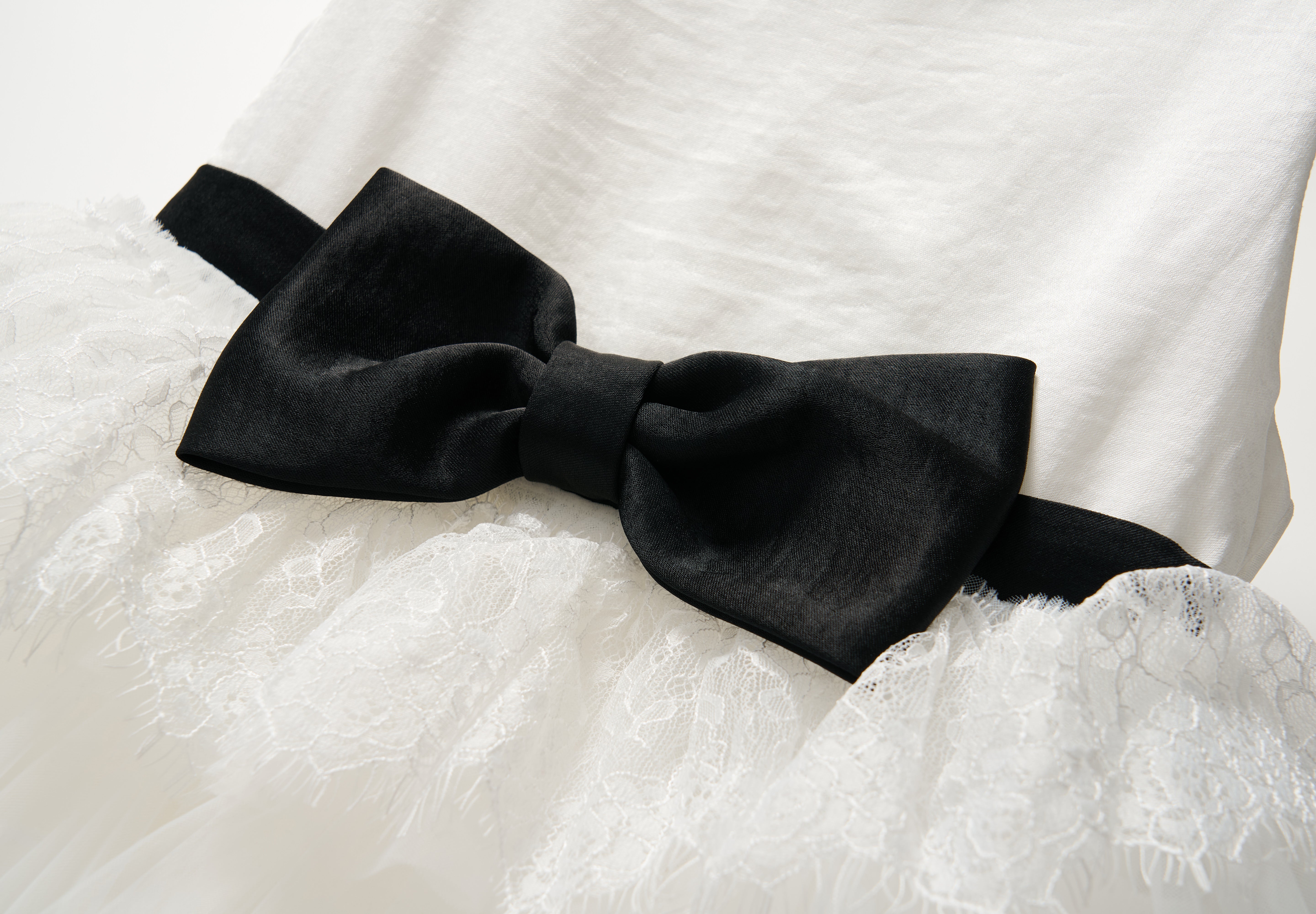 The Monochrome Whisper Dress by KIKI and JOJO features white lace ruffles, a large black velvet waist bow, and a softly layered tulle cake skirt, shown against a plain background.