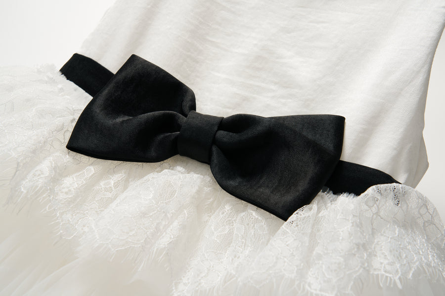 The Monochrome Whisper Dress by KIKI and JOJO features white lace ruffles, a large black velvet waist bow, and a softly layered tulle cake skirt, shown against a plain background.