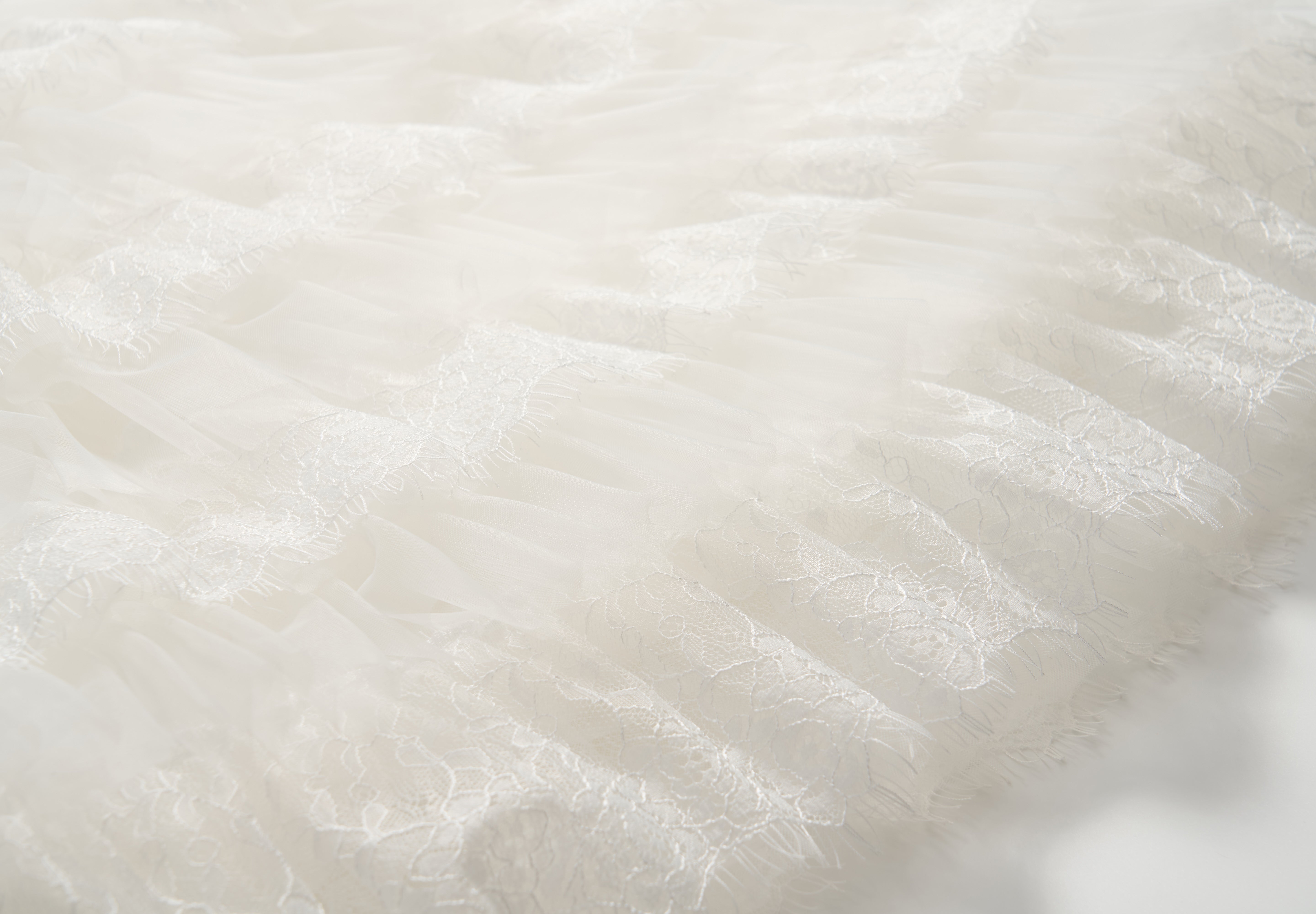 Close-up of the Monochrome Whisper Dress by KIKI and JOJO, featuring delicate white lace and tulle fabric with a textured, layered pattern reminiscent of elegant ruffles.