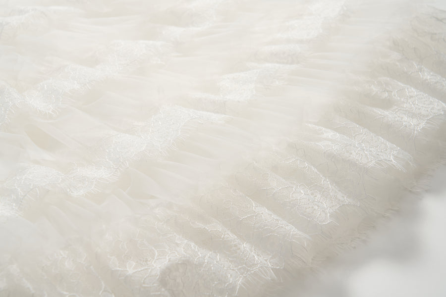 Close-up of the Monochrome Whisper Dress by KIKI and JOJO, featuring delicate white lace and tulle fabric with a textured, layered pattern reminiscent of elegant ruffles.