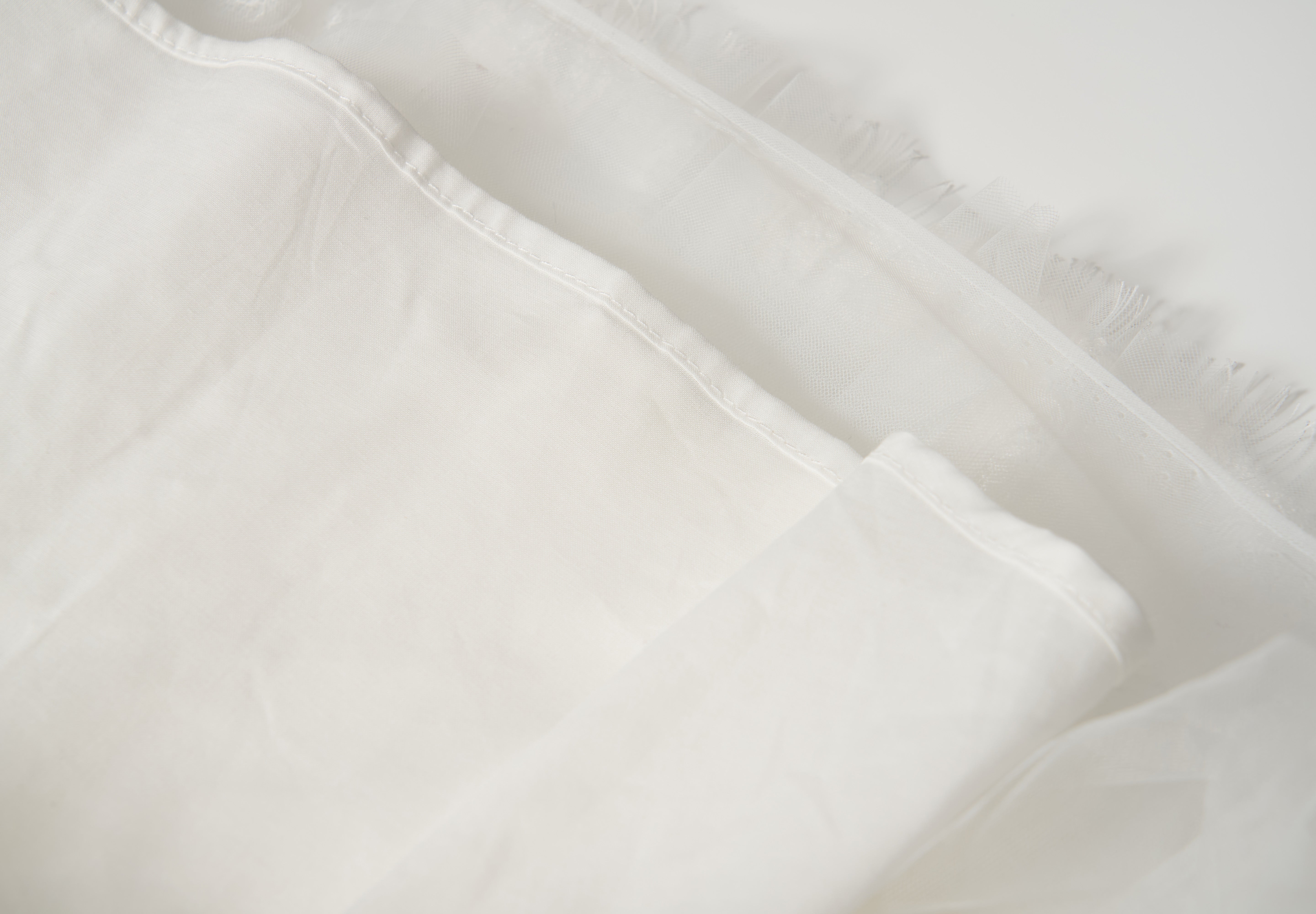 Close-up of the Monochrome Whisper Dress by KIKI and JOJO, showcasing its white, semi-transparent fabric with frayed edge—evoking delicate tulle layers—set against a light background.