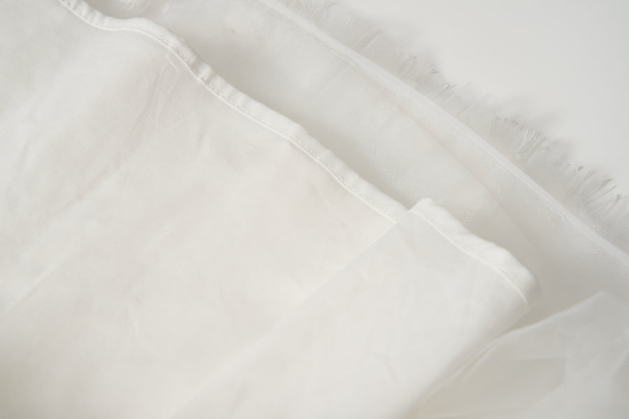 Close-up of the Monochrome Whisper Dress by KIKI and JOJO, showcasing its white, semi-transparent fabric with frayed edge—evoking delicate tulle layers—set against a light background.