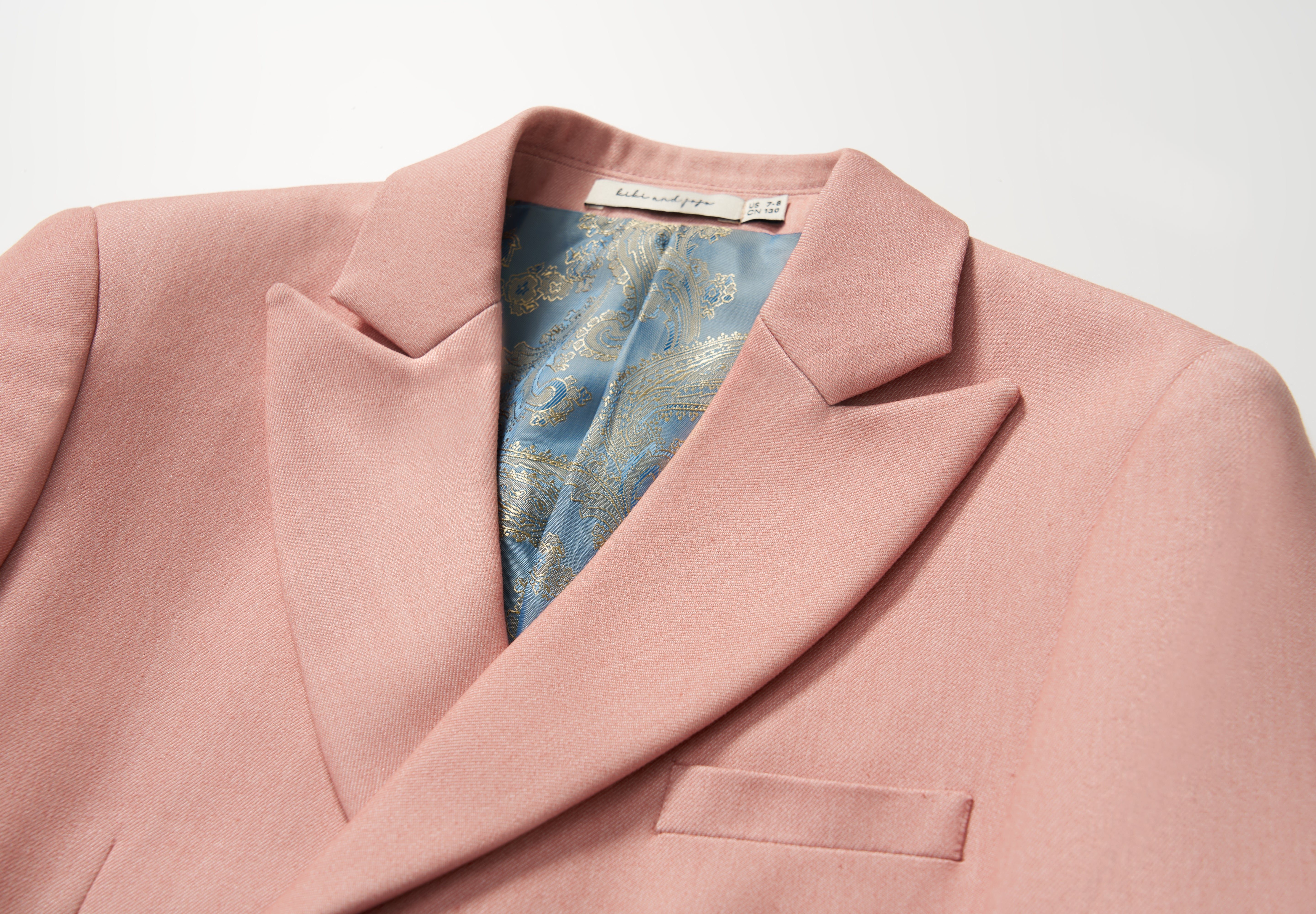 The KIKI and JOJO Blush Reverie Suit Jacket is a blush pink boys’ double-breasted blazer with a notched lapel and blue-gold patterned inner lining, displayed on a white background—ideal for special occasions.
