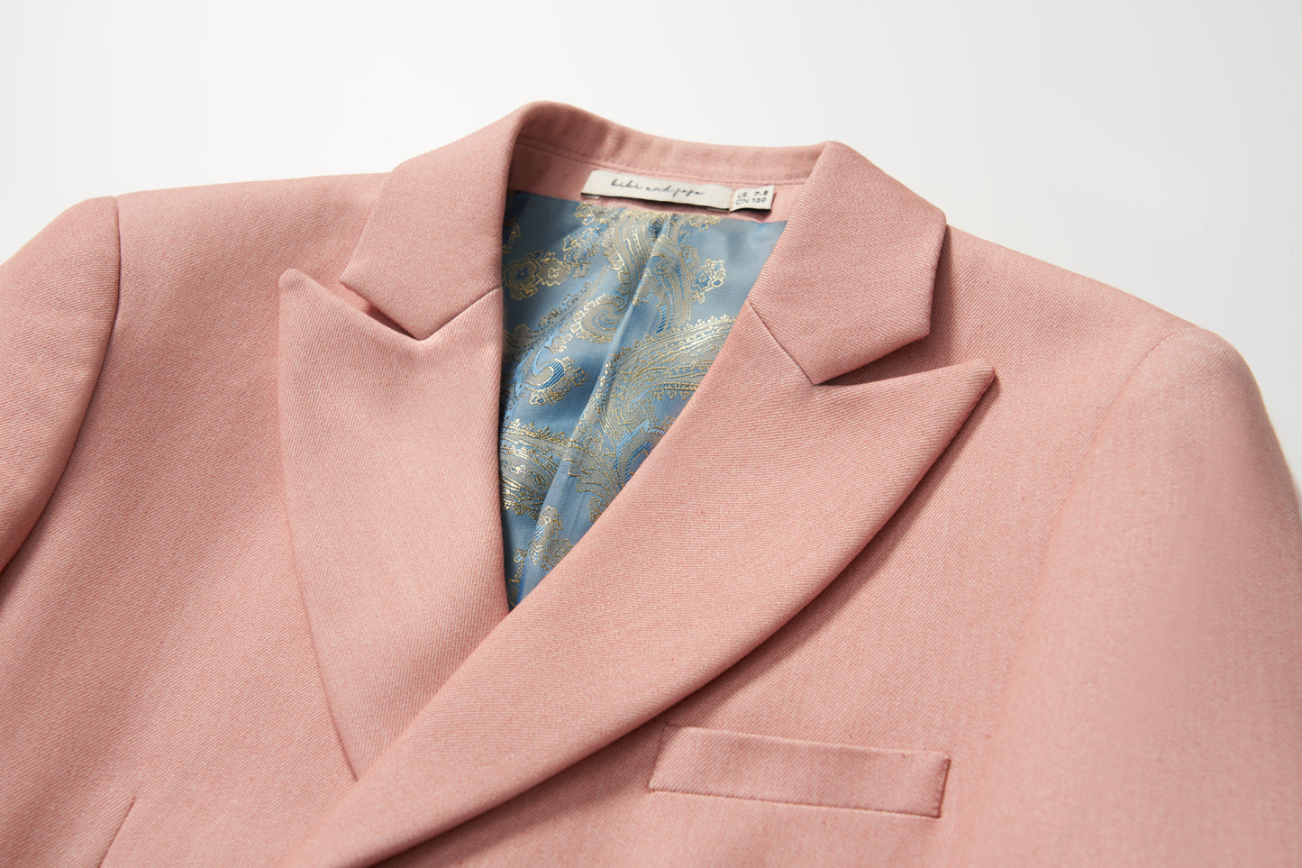 The KIKI and JOJO Blush Reverie Suit Jacket is a blush pink boys’ double-breasted blazer with a notched lapel and blue-gold patterned inner lining, displayed on a white background—ideal for special occasions.