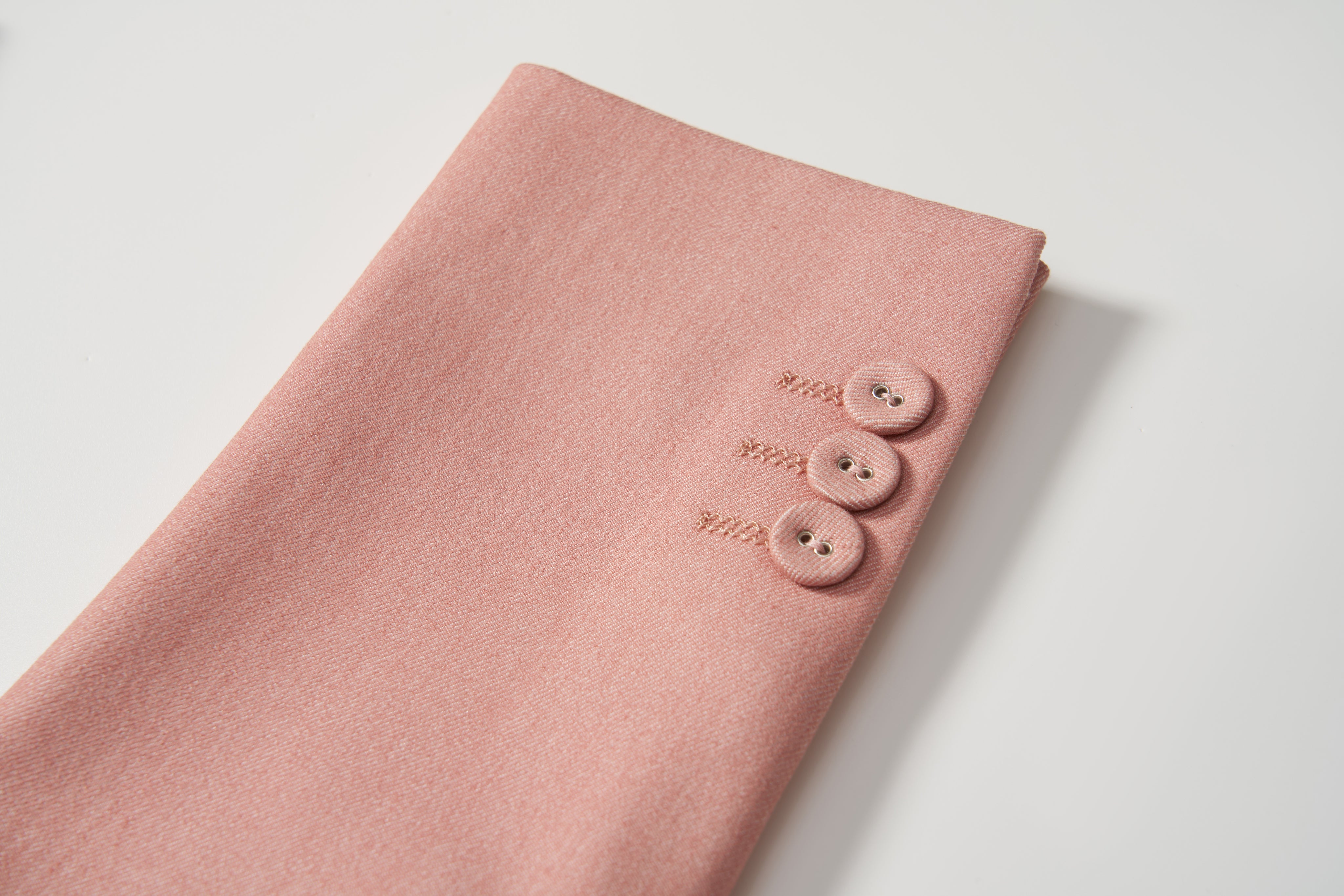 A pair of Blush Reverie Suit Pants by KIKI and JOJO, featuring an adjustable waistband and three matching buttons in a row, is displayed folded on a white surface.