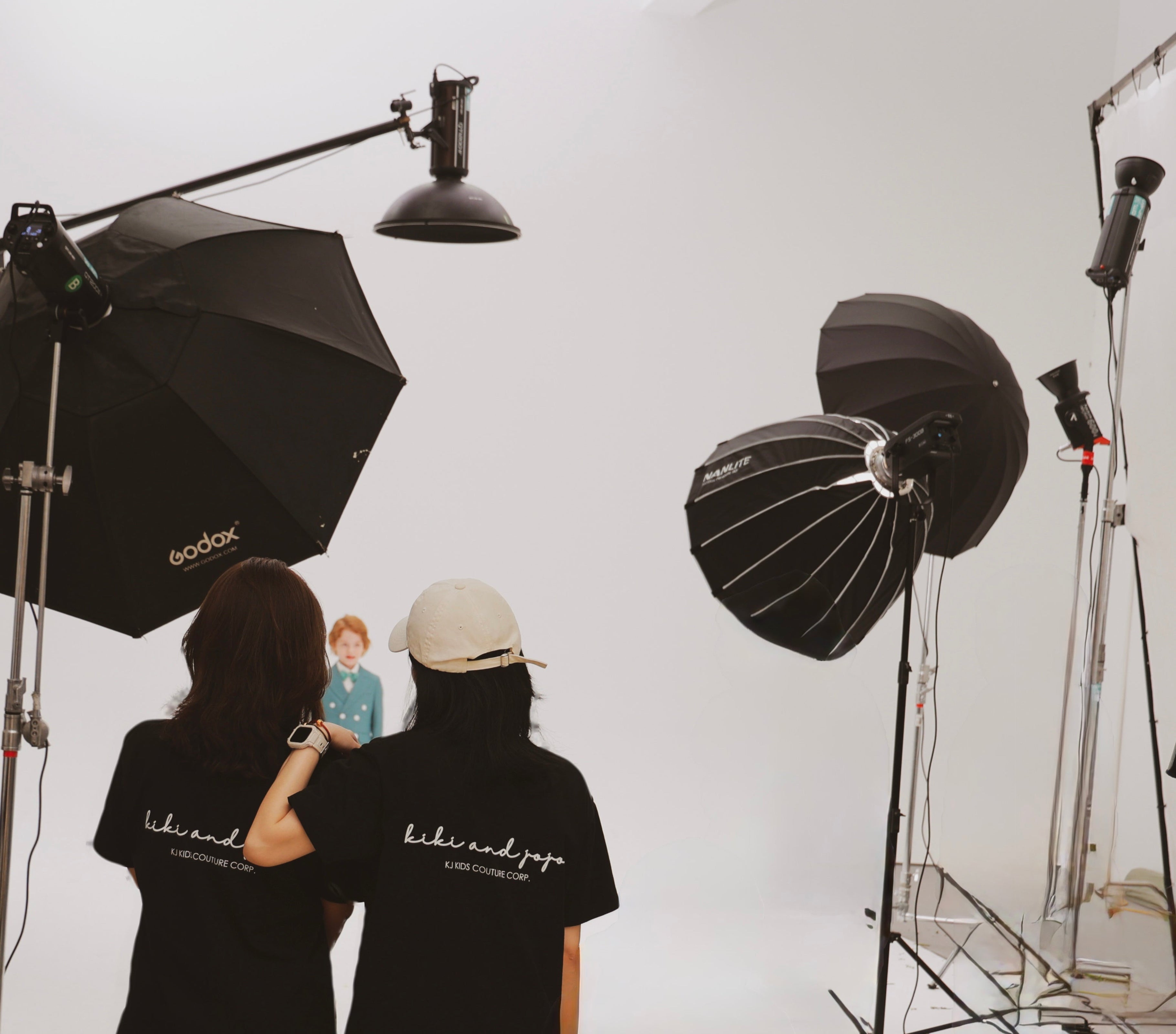 Two people in black shirts stand in a photo studio, facing a model on a white backdrop, surrounded by professional lighting equipment.