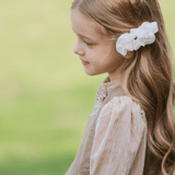 A young girl with long, wavy hair wears the KIKI and JOJO Girls Chiffon Hair Clip in Ivory while standing outdoors in a light beige dress, facing sideways.