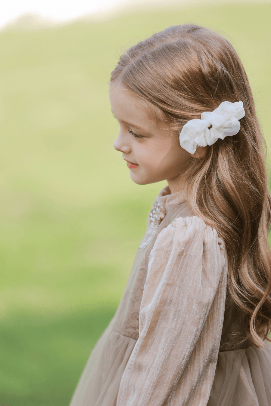 A young girl with long, wavy hair wears the KIKI and JOJO Girls Chiffon Hair Clip in Ivory while standing outdoors in a light beige dress, facing sideways.