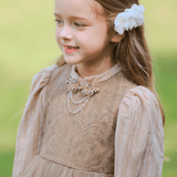 A young girl with long brown hair, wearing a beige dress and the KIKI and JOJO Girls Chiffon Hair Clip in Ivory—a charming, handmade accessory—stands outdoors on a sunny day.