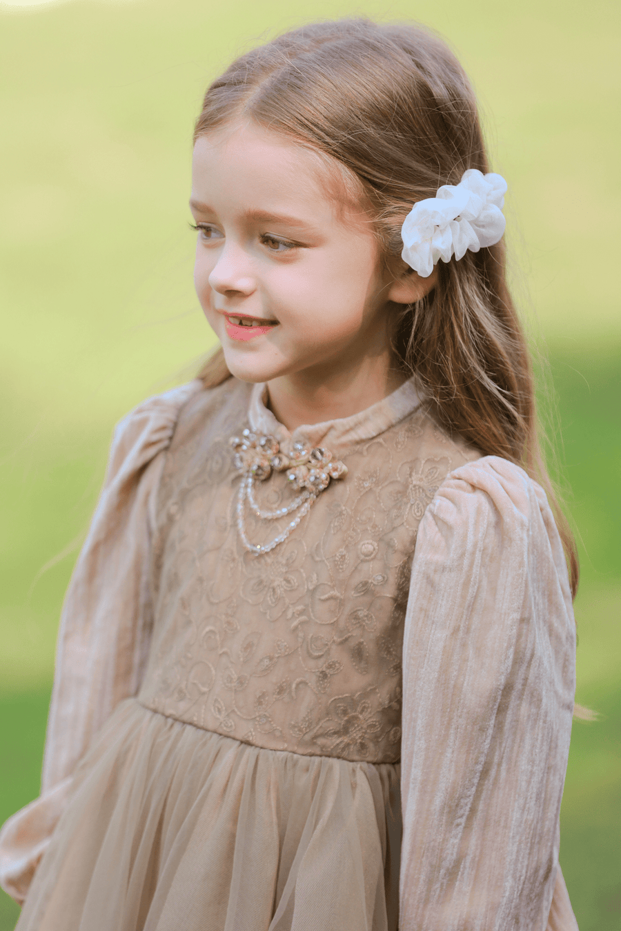 A young girl with long brown hair, wearing a beige dress and the KIKI and JOJO Girls Chiffon Hair Clip in Ivory—a charming, handmade accessory—stands outdoors on a sunny day.