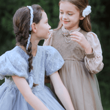 Two young girls in vintage dresses stand outdoors, smiling at each other, each wearing the KIKI and JOJO Girls Chiffon Hair Clip in Ivory, surrounded by lush greenery.