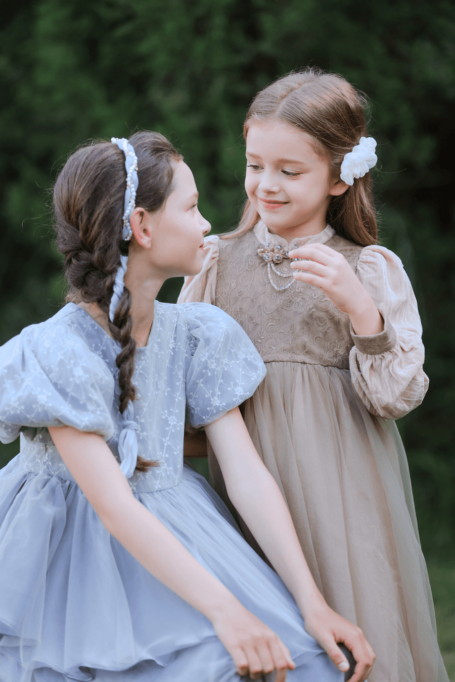 Two young girls in vintage dresses stand outdoors, smiling at each other, each wearing the KIKI and JOJO Girls Chiffon Hair Clip in Ivory, surrounded by lush greenery.