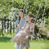 Two young girls in dresses enjoy the outdoors; one sits on a wooden swing while the other stands behind, holding the ropes. The KIKI and JOJO Girls Chiffon Hair Clip - Ivory adds enchanting charm amid the sunlit greenery.
