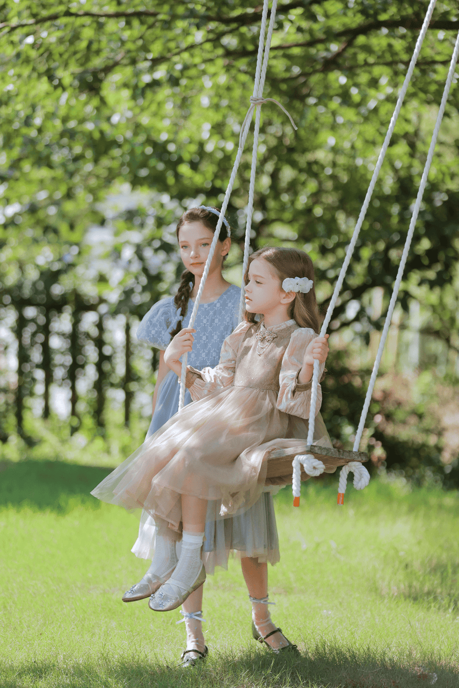 Two young girls in dresses enjoy the outdoors; one sits on a wooden swing while the other stands behind, holding the ropes. The KIKI and JOJO Girls Chiffon Hair Clip - Ivory adds enchanting charm amid the sunlit greenery.
