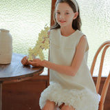 A young girl in a white dress sits on a chair by a wooden table, holding yellow flowers and wearing the KIKI and JOJO Girls Diamanté Padded Hairband in Ivory.