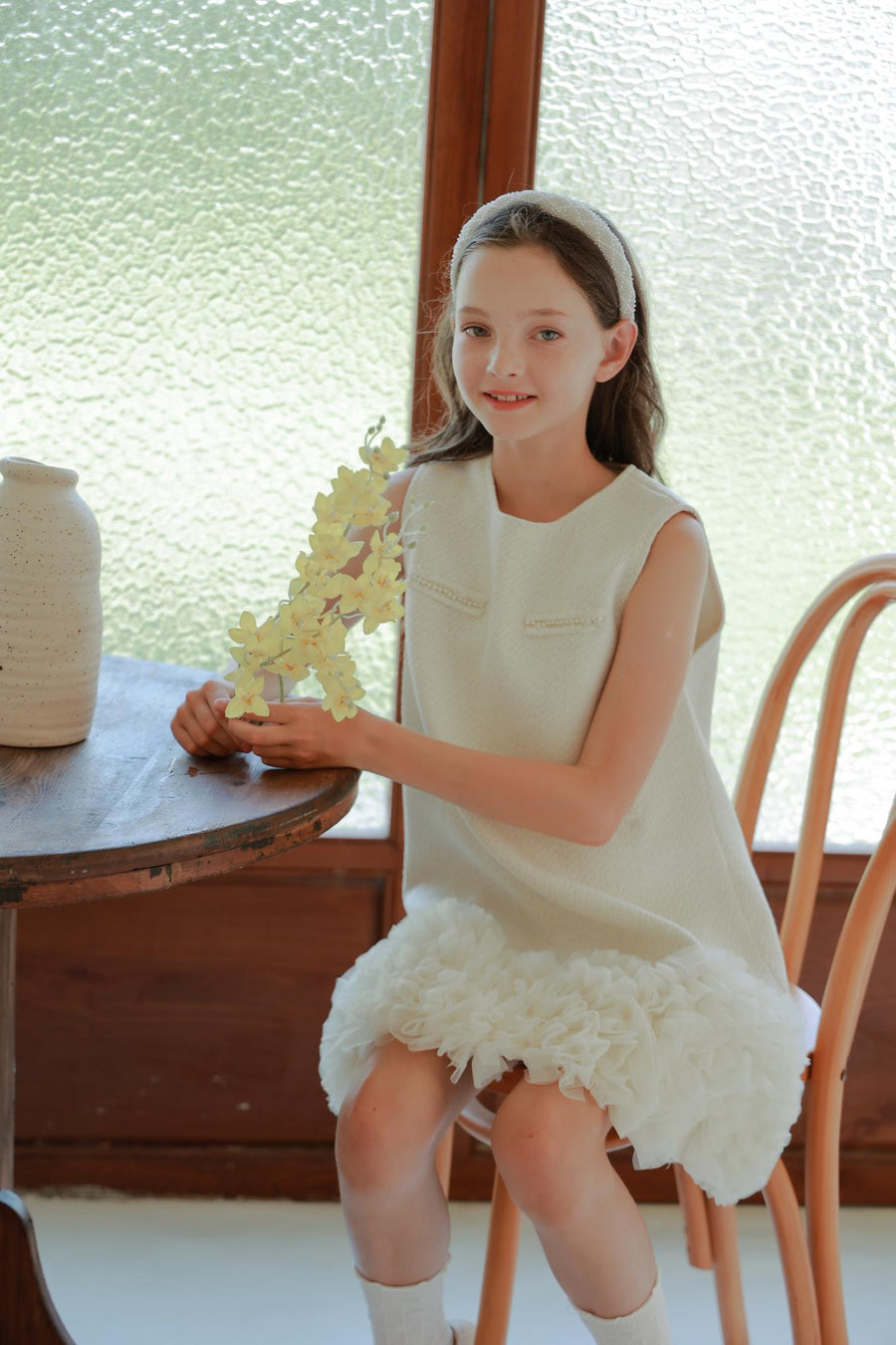 A young girl in a white dress sits on a chair by a wooden table, holding yellow flowers and wearing the KIKI and JOJO Girls Diamanté Padded Hairband in Ivory.