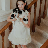 A young girl in a white dress wears the KIKI and JOJO Girls Diamanté Padded Hairband in Ivory as she stands on a staircase holding two small black-and-white puppies, looking at the camera.