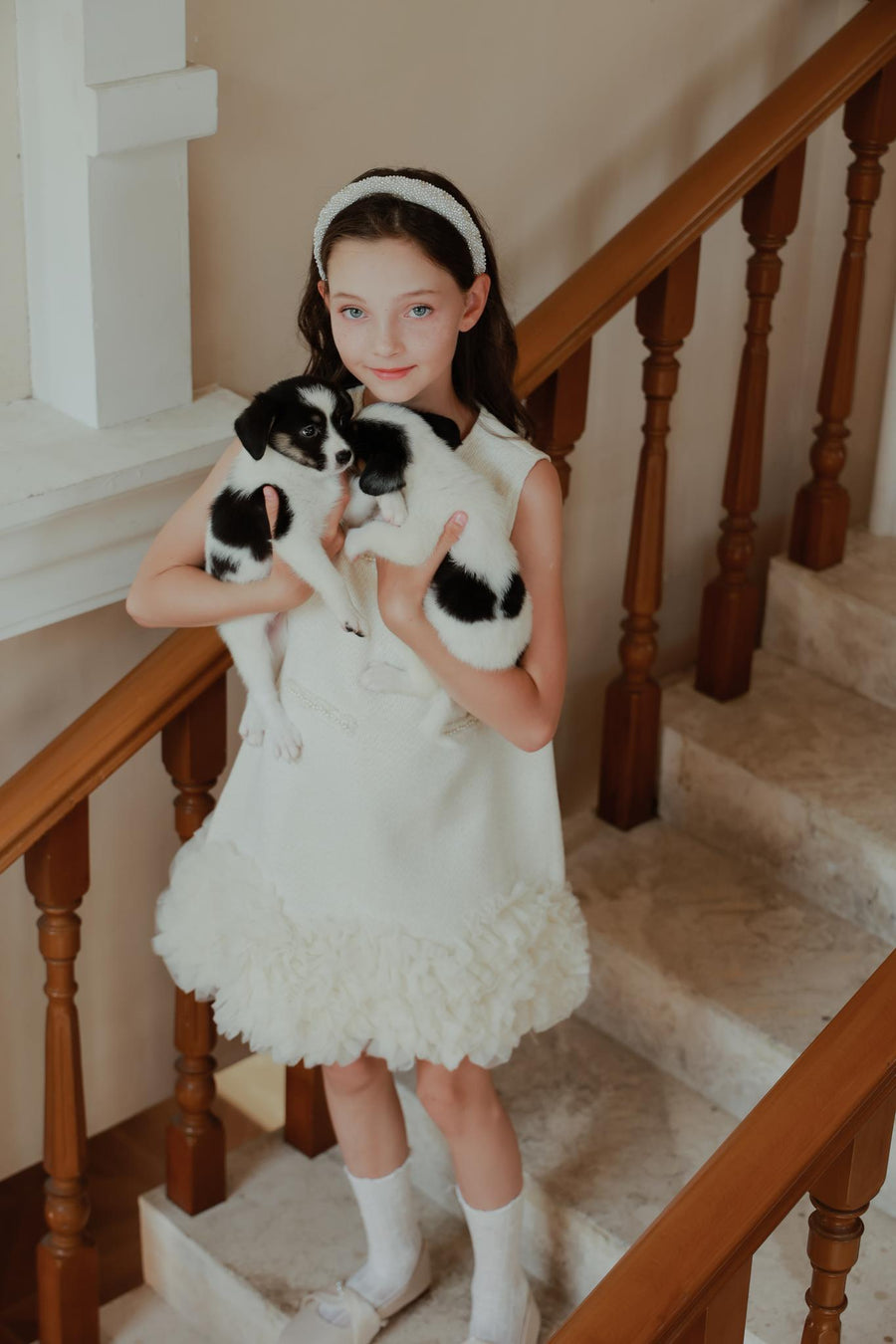 A young girl in a white dress wears the KIKI and JOJO Girls Diamanté Padded Hairband in Ivory as she stands on a staircase holding two small black-and-white puppies, looking at the camera.