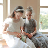 Two young girls in light dresses sit by a sunlit window reading books. One wears the KIKI and JOJO Girls Diamanté Padded Hairband in Ivory. Behind them, a vase with a single wilted flower rests on the windowsill.