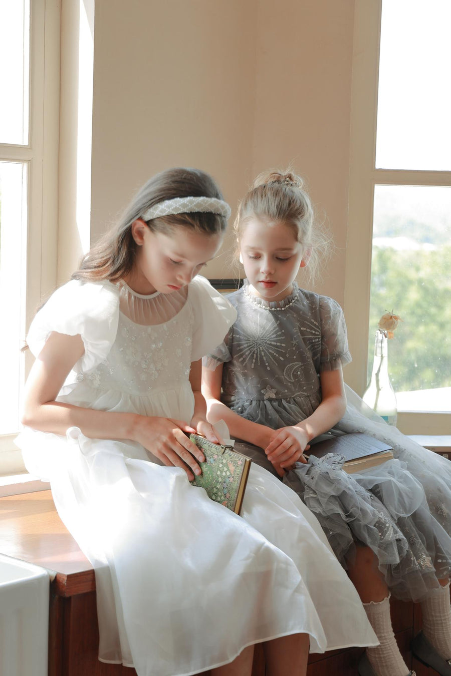 Two young girls in light dresses sit by a sunlit window reading books. One wears the KIKI and JOJO Girls Diamanté Padded Hairband in Ivory. Behind them, a vase with a single wilted flower rests on the windowsill.