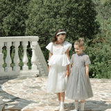 Two young girls in formal dresses walk down stone steps outdoors, one wearing the KIKI and JOJO Girls Diamanté Padded Hairband in Ivory, surrounded by greenery and a white balustrade.
