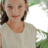 A young girl with light eyes and brown hair wears the KIKI and JOJO Girls Diamanté Padded Hairband in ivory with a sleeveless top, standing before green plants.