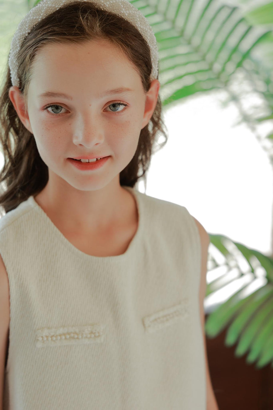 A young girl with light eyes and brown hair wears the KIKI and JOJO Girls Diamanté Padded Hairband in ivory with a sleeveless top, standing before green plants.