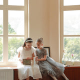 Two young girls in formal dresses sit on a wooden cabinet by large windows—one reads a book while the other, wearing the KIKI and JOJO Girls Diamanté Padded Hairband in Ivory, looks on.