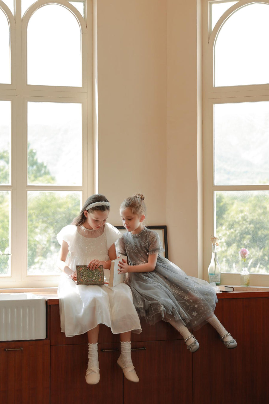 Two young girls in formal dresses sit on a wooden cabinet by large windows—one reads a book while the other, wearing the KIKI and JOJO Girls Diamanté Padded Hairband in Ivory, looks on.