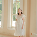 A young girl stands indoors near large arched windows, sunlight streaming in, wearing a white dress and the KIKI and JOJO Girls Diamanté Padded Hairband in Ivory.