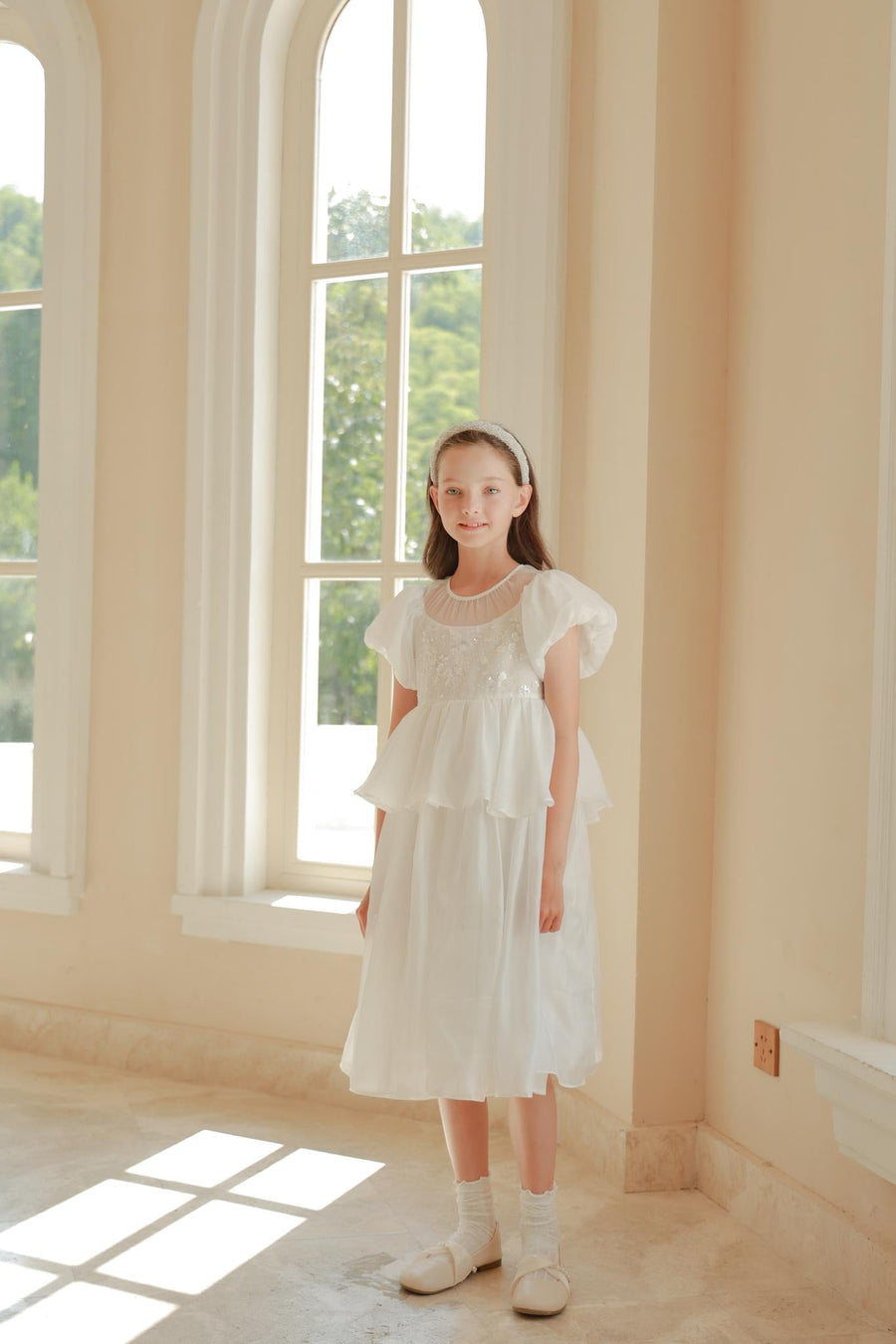 A young girl stands indoors near large arched windows, sunlight streaming in, wearing a white dress and the KIKI and JOJO Girls Diamanté Padded Hairband in Ivory.