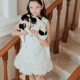 A girl wearing the KIKI and JOJO Girls Elegance A-Line Chiffon Trim Dress in Champagne stands on a staircase, holding two black and white puppies, and looks at the camera.