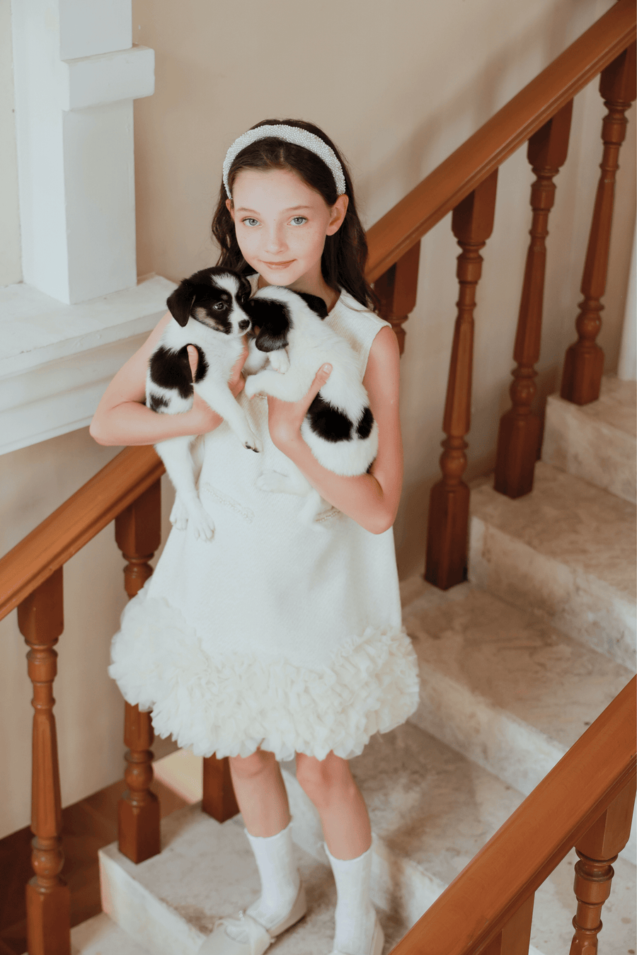 A girl wearing the KIKI and JOJO Girls Elegance A-Line Chiffon Trim Dress in Champagne stands on a staircase, holding two black and white puppies, and looks at the camera.