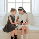Two young girls sit by a window, wearing KIKI and JOJO Girls Elegance A-Line Chiffon Trim Dress in champagne, gently holding and petting a small black and white puppy on their laps.