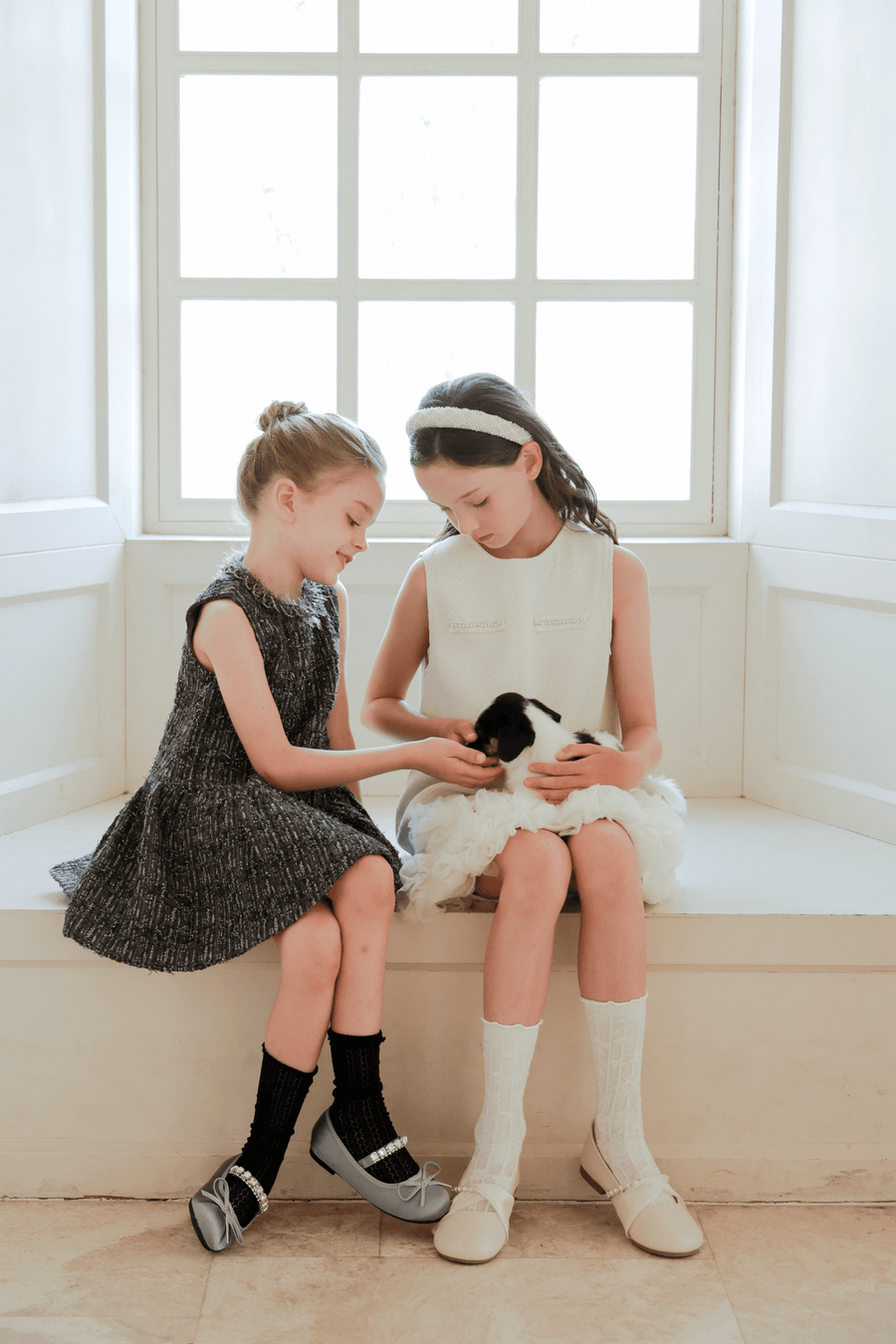 Two young girls sit by a window, wearing KIKI and JOJO Girls Elegance A-Line Chiffon Trim Dress in champagne, gently holding and petting a small black and white puppy on their laps.