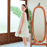 A girl wearing the KIKI and JOJO Girls Elegance A-Line Chiffon Trim Dress in Champagne stands indoors by a wooden full-length mirror, with glass doors and green leaves visible in the background.