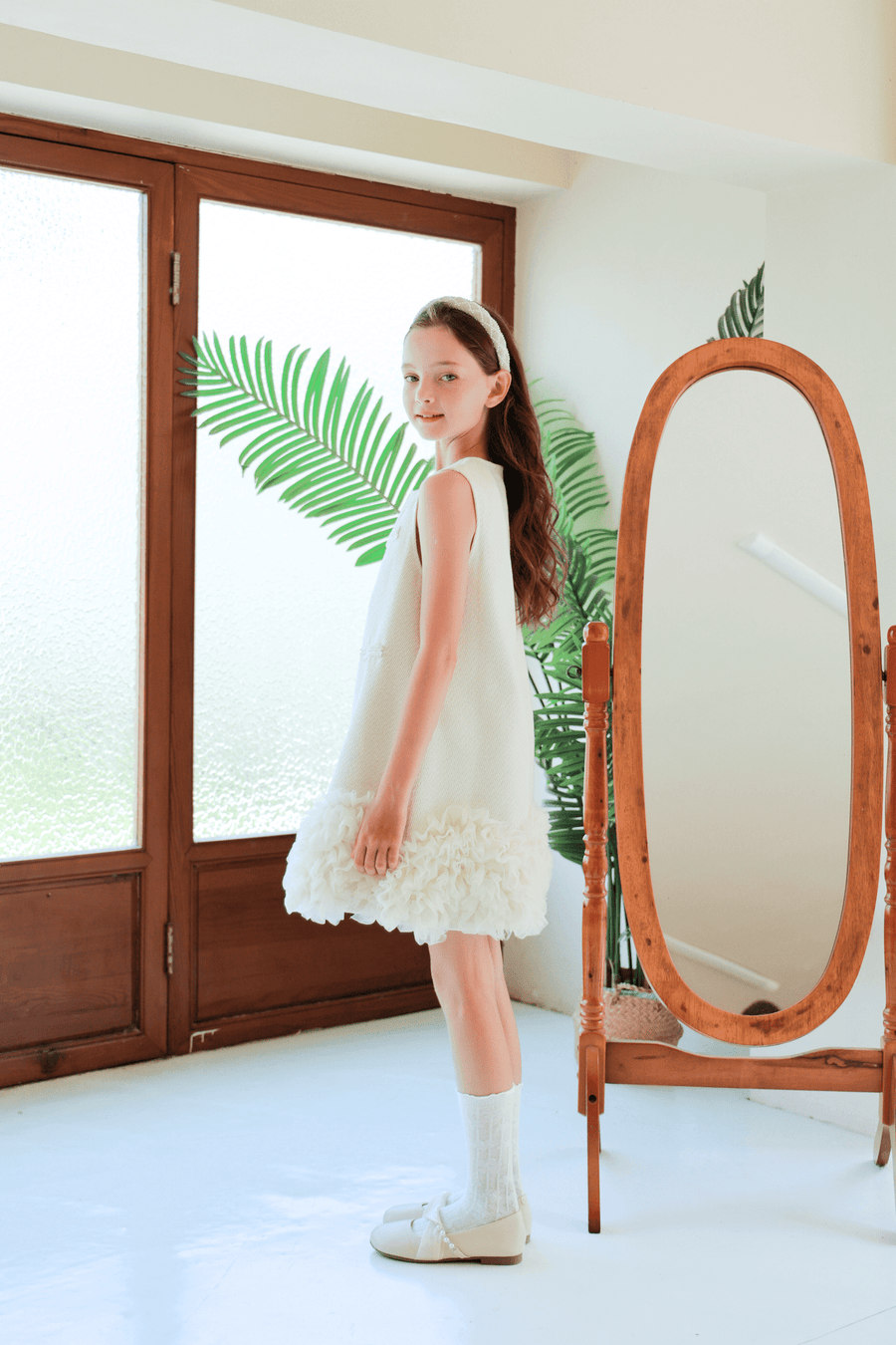 A girl wearing the KIKI and JOJO Girls Elegance A-Line Chiffon Trim Dress in Champagne stands indoors by a wooden full-length mirror, with glass doors and green leaves visible in the background.